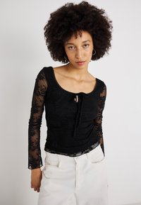 Μαύρη μπλούζα με μακριά μανίκια και λουλουδάτη δαντέλα, με στρογγυλή λαιμόκοψη και ζώνη μπροστά. Συνδυασμένη με λευκά παντελόνια ψηλής μέσης.