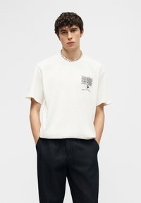 Νέος άνδρας με σγουρά μαλλιά, φορώντας λευκό μπλουζάκι με στάμπα και σκούρο παντελόνι, στέκεται με τα χέρια στις τσέπες μπροστά σε απλό φόντο.