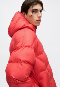 Joven con cabello oscuro que lleva una chaqueta acolchada roja brillante con capucha, mirando por encima del hombro contra un fondo claro y liso.