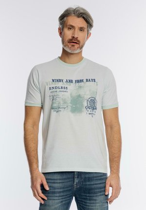 Mężczyzna z siwymi włosami i brodą, ubrany w jasnografitowy zielony t-shirt z nadrukiem oraz niebieskie dżinsy, stojący na białym tle.