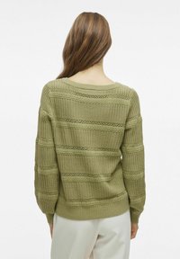 Oliwkowy sweter z dzianiny o luźnym kroju, z teksturowanymi poziomymi paskami i skomplikowanymi wzorami przypominającymi koronkę wzdłuż szwów.