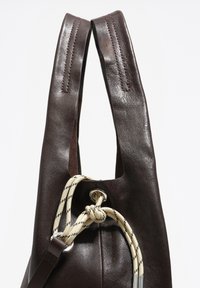 Brązowa skórzana torba z grubymi, wyraźnymi uchwytami. Posiada zapięcie na sznurek z kremowego sznura z czarnymi paskami. Gładka tekstura.