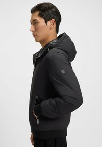 Man som bär en svart huvtröja med dragkedjefickor, stående i profil med en hand i fickan mot en enkel ljus bakgrund.