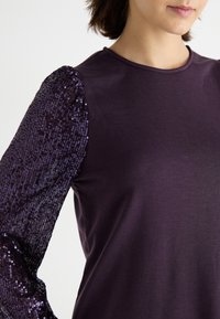 Lila skjorta med solid kropp och paljettsprutad ärm. Mjuk tyg med glittrande, texturerade paljetter på en ärm, vilket skapar en kontrasterande design.