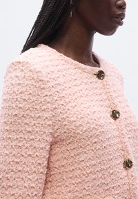 Rózsaszín tweed zakó, kerek nyakkivágással, texturált felülettel és három arany gombbal díszítve.