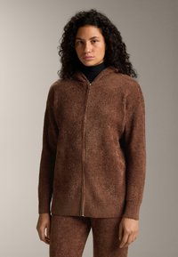 Hnedý mikinový sveter s kapucňou a zipsom vpredu, vyrobený z textúrovaného materiálu, s dlhými rukávmi a voľným strihom. Má strieborný zips a jemný, chlpatý povrch.