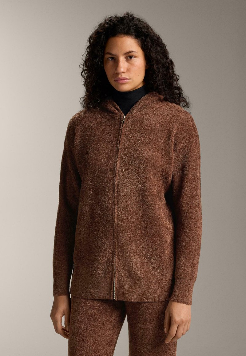 Hnedý mikinový sveter s kapucňou a zipsom vpredu, vyrobený z textúrovaného materiálu, s dlhými rukávmi a voľným strihom. Má strieborný zips a jemný, chlpatý povrch.