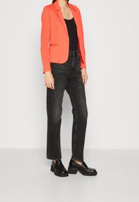 Blazer ajusté corail porté sur un haut noir, associé à un jean droit noir taille haute et des chaussures noires épaisses. Lignes épurées et design moderne.