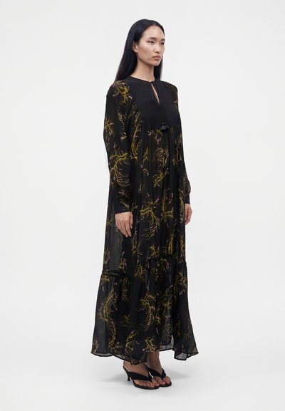 Femme debout portant une longue robe noire transparente avec des motifs floraux jaunes et roses et des sandales noires à talons sur un fond blanc uni.