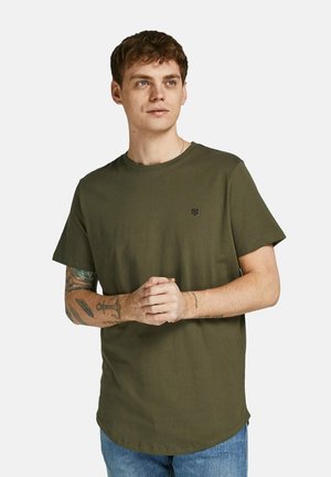 Young man with brown hair wearing olive green short-sleeve t-shirt and blue jeans, hands clasped, looking slightly right, with tattoos on left arm.