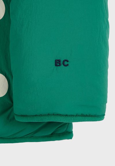 Grön quiltad jacka med stora vita prickar. Har en broderad "BC"-logotyp i marinblått på ärmen. Slät textur och rundad form.