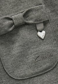 Grijze stof met een gladde textuur heeft een strikdetail en een hartvormige metalen charme gegrafeerd met "Clarks." Het logo is geborduurd.