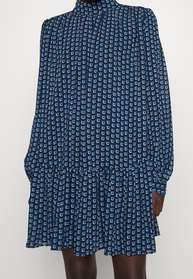 Robe bleue à manches longues avec motif carré bleu clair, col montant, jupe froncée et poignets ajustés sur un mannequin à la peau foncée.