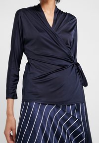 Námornícko modrý zavinovací top vyrobený z hladké, elastické látky s dlouhými rukávy a zavazováním v oblasti pasu, zkombinovaný se pruhovanou sukní pro kontrast.