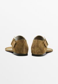 Brune ruskindsandaler med flad sål, bagrem og en dekorativ sløjfe på siden. Blød tekstur og minimalistisk design.