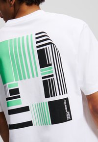 Valkoinen lyhythihainen t-paita, jossa on vihreä- ja mustasävyinen geometrinen graafinen kuvasto selkäpuolella, ja jossa on brändin nimi "Karl Lagerfeld Jeans."