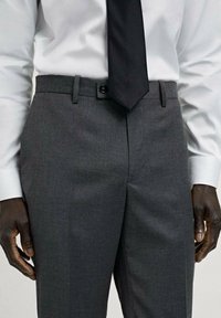 Pantaloni de costum gri, confecționați dintr-un material Neted, cu față plată, buzunare laterale și bucle pentru curea, asortați cu o cămașă albă și o cravată neagră.