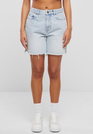 Kvinde iført lyseblå højtaljede denimshorts med frynser, hvide sokker og hvide sneakers mod en ensfarvet baggrund.