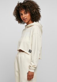 Sweat à capuche court en crème, fabriqué dans un tissu doux, avec une capuche, arborant un grand logo blanc et un détail de tag noir sur le côté. Associé à un jogger assorti.