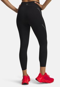 Zwarte high-waisted legging met een gladde textuur, een aansluitend ontwerp, reflecterende zilveren accenten en geperforeerde details, gecombineerd met felrode sneakers.