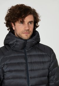 Svart puffjacka med hög krage, med en quiltad design. Dragkedjestängning, lätt material och subtil varumärkeslogotyp på bröstet.