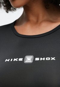 Čierny atletický top s okrúhlym výstrihom, na hrudi s výrazným bielym logom "NIKE SHOX", vyrobený z hladkého, pružného materiálu.