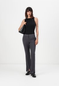 Čierny rebrovaný top bez rukávov, šedé elegantné nohavice s bočnými rozparkami a čierne topánky. Modelka drží čiernu kabelku. Jednoduché biele pozadie.