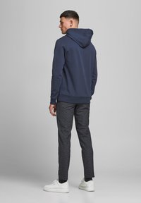 Haut à capuche marine avec cordon de serrage, associé à un pantalon sombre à motifs et des baskets blanches. Le tissu semble lisse et ajusté.