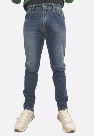 Uomo che indossa jeans slim fit blu e sneakers bianche, in piedi davanti a uno sfondo semplice.