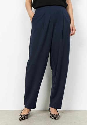 Donna che indossa pantaloni ampi a vita alta blu navy con pieghe, top nero e tacchi a punta con stampa a zebre, in piedi contro un muro bianco.
