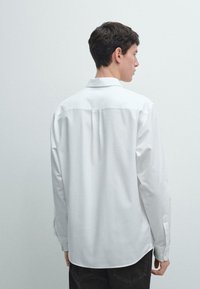Camicia bianca a maniche lunghe con colletto abbottonato, tessuto liscio, cucitura sul dietro del giogo e orlo curvo, abbinata a pantaloni scuri.