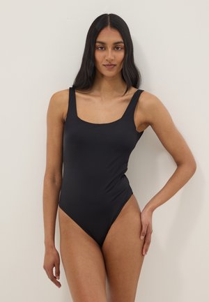 Femme aux longs cheveux foncés portant un body noir moulant sans manches, debout avec une main sur la hanche sur un fond uni.