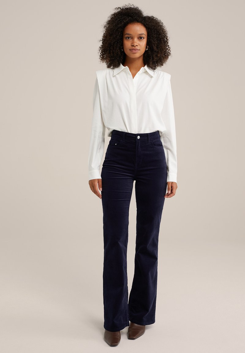 Camisa blanca de manga larga con detalles en los hombros, combinada con pantalones acampanados de pana azul marino de cintura alta. Zapatos marrones de punta.