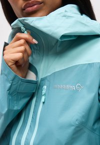 Inimene tõmbab lukku helesinist vetthülgavat jopet kõrge kaeluse ja nähtava NORRØNA logoga rinnal.