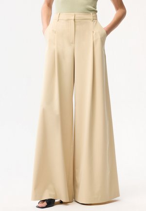 Beige hoch taillierte Hosen mit weitem Bein und Frontfalten, seitlichen Taschen, getragen mit schwarzen offenen Sandalen und einem hellgrünen ärmellosen Oberteil.