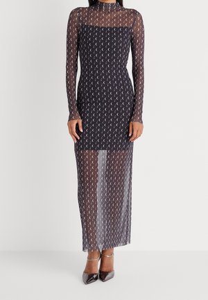 Frau trägt ein durchsichtiges schwarzes Maxikleid mit langen Ärmeln, verziert mit Mondsichelmustern, und schwarze Absatzschuhe mit Knöchelriemen.