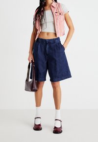 Shorts de mezclilla con un corte relajado, que presentan una cintura alta, un color azul profundo y costuras visibles. Combinados con un chaleco corto de rayas rosas y zapatos oscuros.