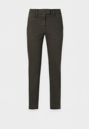 Femme aux longs cheveux foncés portant une chemise blanche à manches courtes boutonnée, un pantalon slim foncé et des baskets blanches, debout les mains dans les poches.