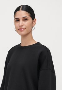 Jovem mulher com cabelo escuro e liso para trás usa argolas de prata e um sweatshirt preto simples, olhando suavemente para a câmara.
