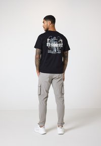 Camiseta negra con un diseño gráfico que incluye la palabra "resiliente" en la parte posterior, combinada con pantalones cargo gris claro y zapatillas blancas.