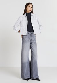 Camicia a righe bianche e blu navy con bottoni, abbinata a jeans grigi a vita alta e a gamba larga. La modella indossa un dolcevita scuro sotto.