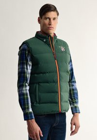 Chaleco acolchado verde con cuello alto, que presenta una cremallera frontal con detalles en rojo y amarillo, y un logo bordado en el pecho.
