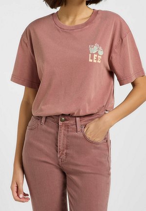 Woman wearing dusty pink Lee t-shirt with floral logo and matching high-waist jeans, one hand in pocket against a plain background.
