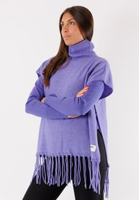 Poncho viola lavorato a maglia con collo alto, caratterizzato da dettagli in frange all'orlo, vestibilità morbida e un'etichetta sul lato. Indossato sopra una maglietta a maniche lunghe.