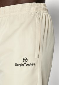 Lys beige treningsshorts laget av teksturert stoff, med elastisk midjebånd og en svart brodert logo nederst til venstre.