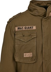 Military-style jacket in olive green, featuring a high collar, multiple pockets, and fabric patches with "M65 GIANT" in black text.