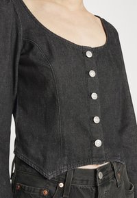 Top cropped en denim noir avec des longues manches, un décolleté carré et six boutons en argent sur le devant ; présente un ourlet effiloché et un tissu texturé.
