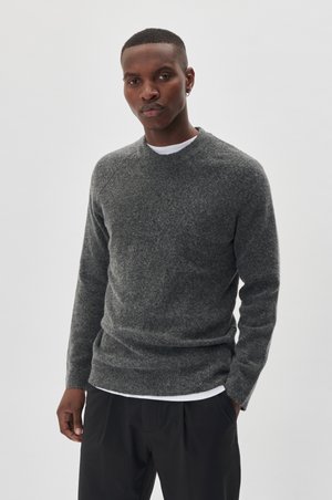 Jersey de lana gris con cuello redondo, puños de canalé y un acabado texturizado, usado sobre una camiseta blanca. El modelo está de pie contra un fondo neutro.