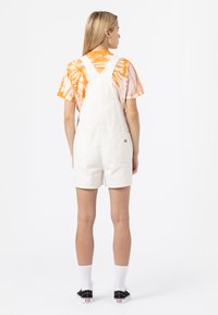 Hvit bomullsoverallsshorts med justerbare skulderstropper, to baklommer og en liten brodert logo. Kombinert med en oransje tie-dye skjorte.