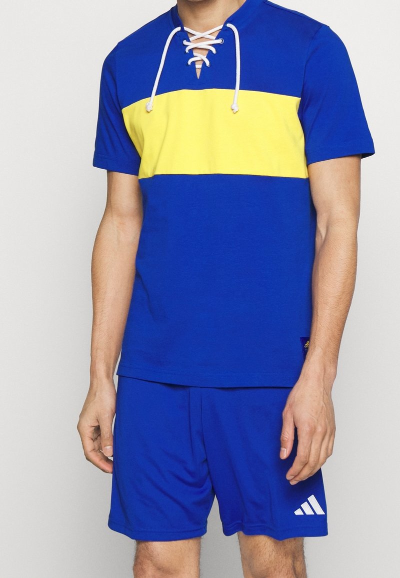 Modré atletické tričko s žlutým vodorovným pruhem, krátkými rukávy a šněrovacím výstřihem, doplněné o odpovídající modré šortky s bílým logem.